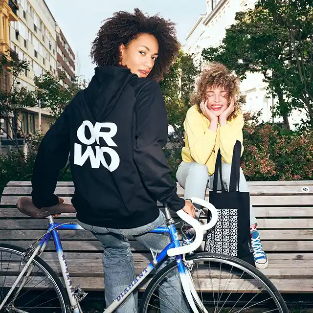 Zwei junge Frauen genießen die frische Luft. Eine sitzt auf einem Fahrrad und trägt einen stylischen ORWO-Hoodie, während die andere auf einer Bank sitzt, lacht und stolz eine ORWO-Stofftasche präsentiert.