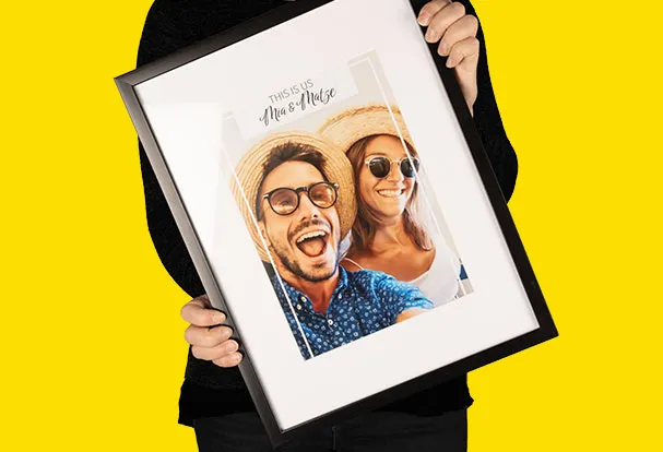 Eine Frau hält stolz einen schwarzen Bilderrahmen mit weißem Passepartout. Das Fotoposter darin zeigt ein glückliches Paar in Urlaubsstimmung.