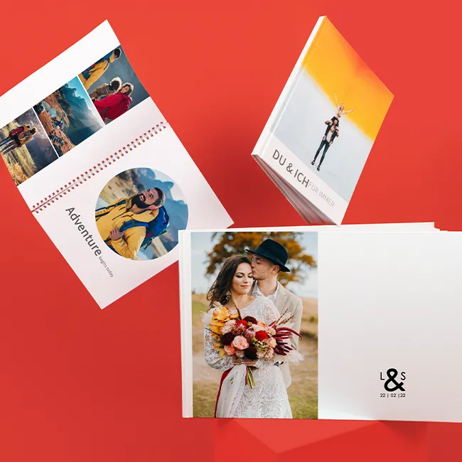 Drei Fotobücher in kreativer Anordnung: Hochzeitsbuch, Wanderbuch mit Ringbindung und Paarbuch „Du & ich für immer“, roter Hintergrund.