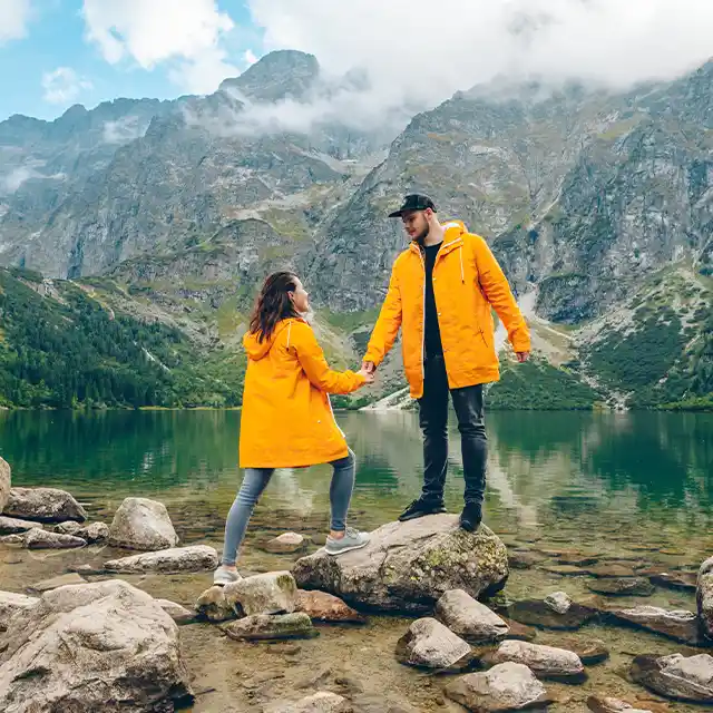 Ein Paar in leuchtend gelben Mänteln steht vor einem kristallklaren Bergsee. Der Mann steht auf einem großen Findling und reicht der Frau die Hand.