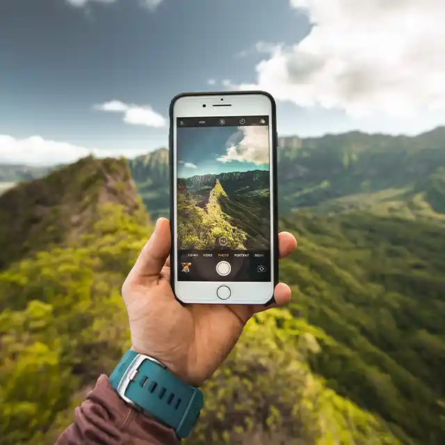 Eine grün bewachsene Berglandschaft im Hintergrund, während ein Wanderer in Nahaufnahme seine Hand mit Smartphone hält und ein Foto macht.