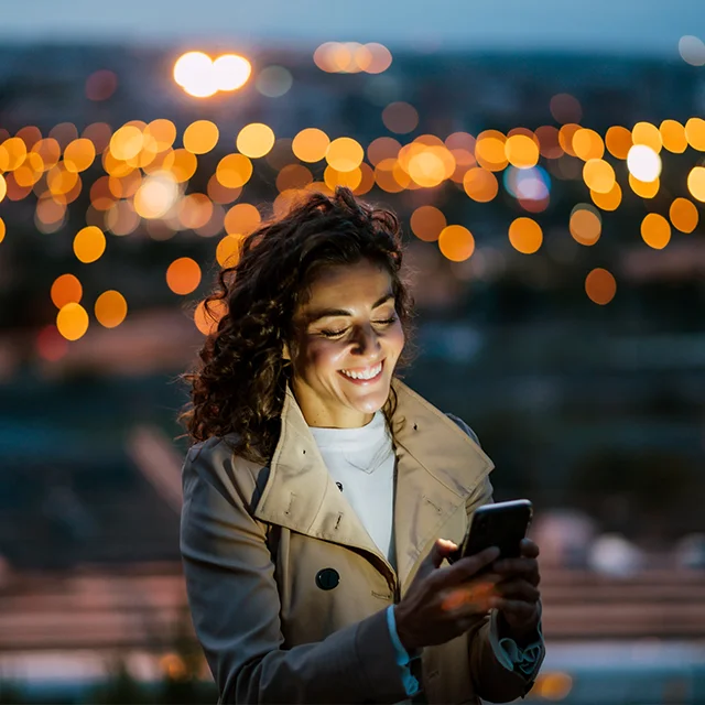 Abendstimmung: Ein Smartphone beleuchtet das Gesicht der im Vordergrund stehenden Person, während der Bokeh-Effekt im Hintergrund für eine traumhafte Atmosphäre sorgt.