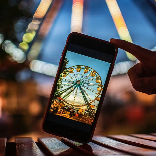 Riesenrad mal anders: Ein Smartphone im Vordergrund fängt das beleuchtete Riesenrad im Hintergrund als Bokeh ein.
