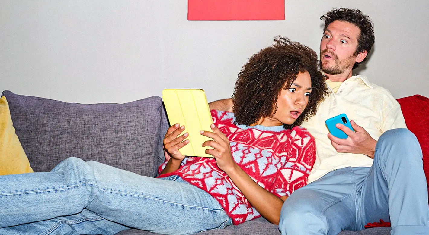 Valentinstag Fotogeschenke mit humorvollem Paarmotiv: Frau und Mann sitzen auf dem Sofa und schauen überrascht auf Tablet und Smartphone