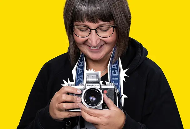 Lächelnde Frau mit Brille hält eine alte Praktica-Kamera vor gelbem Hintergrund. Entdecke die Faszination Analogfotografie mit ORWO.