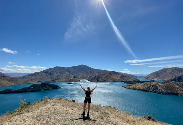 Junge Frau steht mit ausgebreiteten Armen auf einer Klippe mit Blick auf einen tiefblauen See und Berglandschaft unter strahlender Sonne. Lass deine Urlaubsfotos bei ORWO drucken!